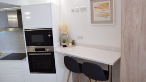 Une cuisine compacte dotée d’un four encastré, d’un four à micro-ondes et d’un plan de travail blanc. On y trouve deux chaises noires et une petite table agrémentée d’objets décoratifs.