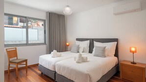 Cama doble con dos almohadas, mesita de noche con lámpara, una silla y una ventana con cortinas.