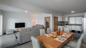 Una sala moderna con un sofá, una mesa de comedor dispuesta con frutas y bebidas, y un televisor.