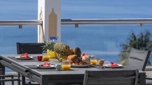 Una mesa dispuesta con alimentos para el desayuno, como frutas, pasteles y jugo de naranja, con vistas a un paisaje marino.