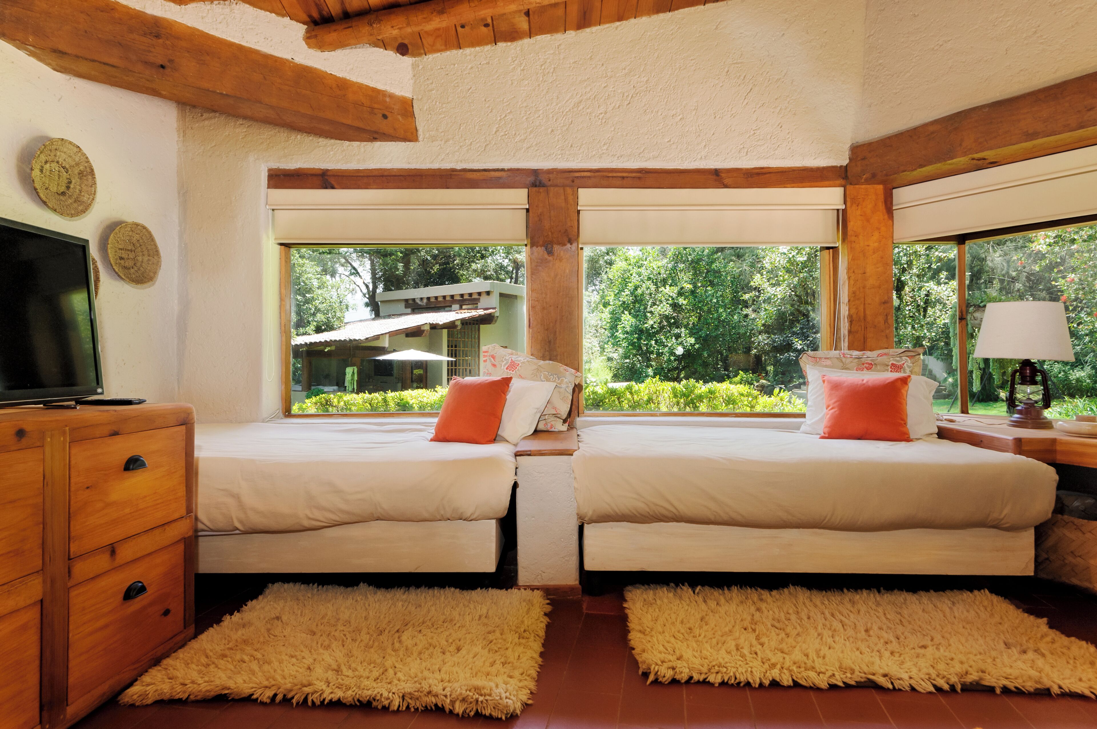 A bedroom with two beds, wooden headboard, large windows, and a TV on a wooden dresser.
