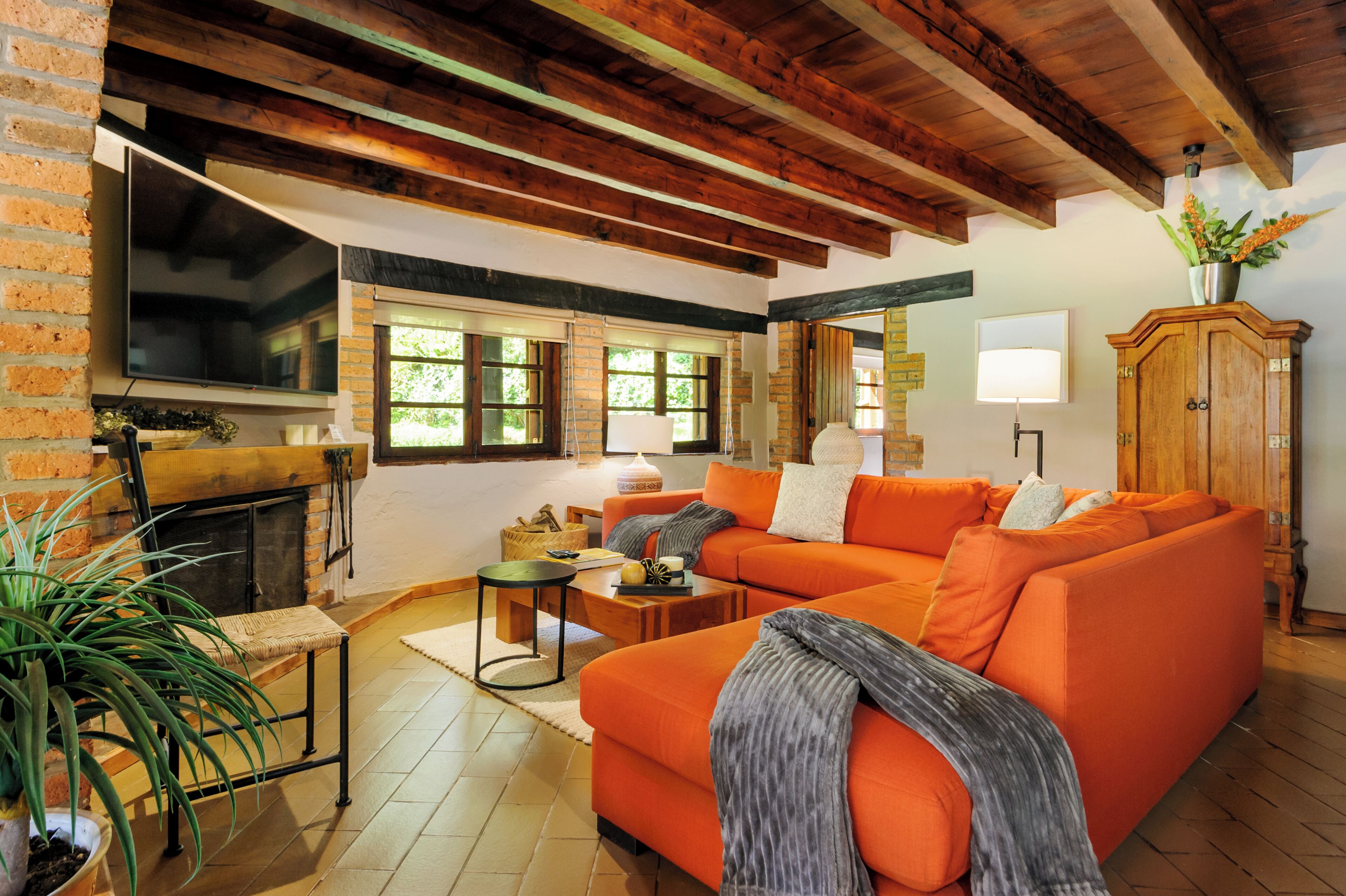 A living room with a brick fireplace, orange sofa, wooden beams, and a large window.