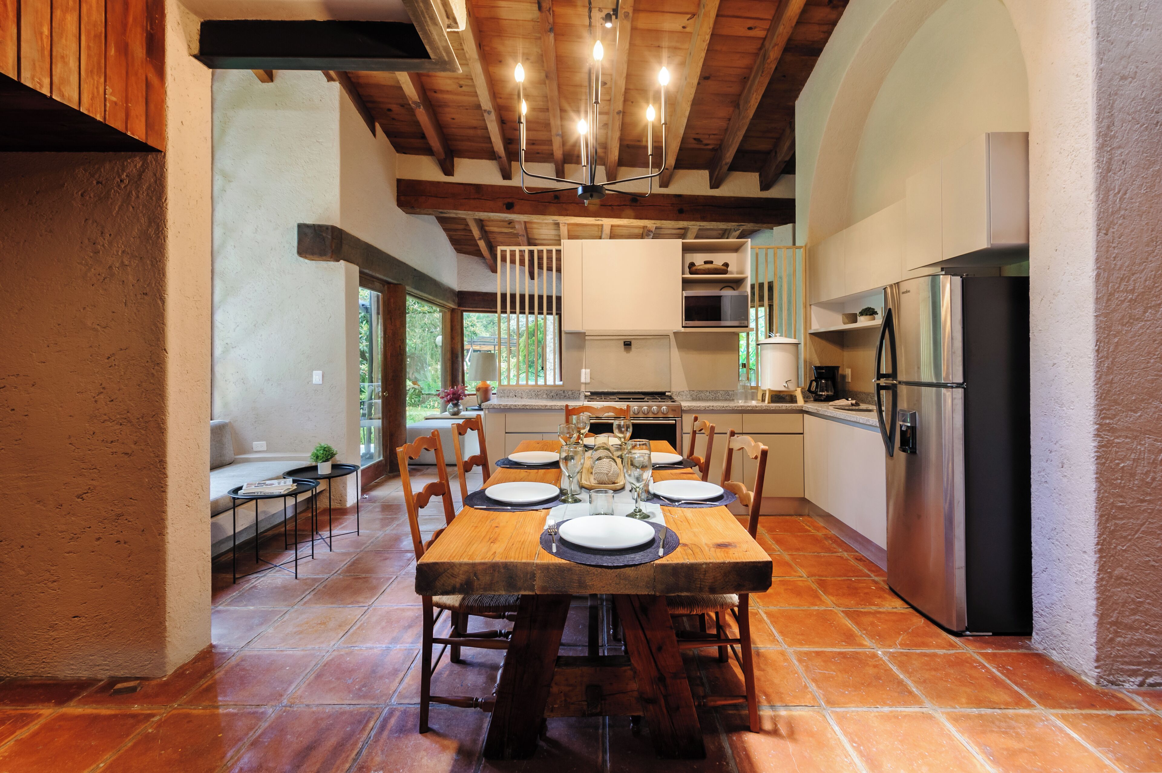 A spacious kitchen with a wooden dining table set for six, a stainless steel refrigerator, and a large window providing natural light.
