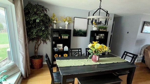 Un coin repas avec une table en bois foncé, une nappe verte et un vase rempli de fleurs jaunes. Des chaises sont disposées autour de la table, ainsi qu’une plante en pot et des étagères ornées d’objets décoratifs.