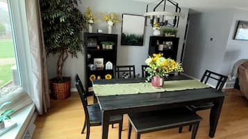 A dining area with a dark wooden table, green tablecloth, and a vase of yellow flowers. There are chairs around the table, a potted plant, and shelves with decorative items.