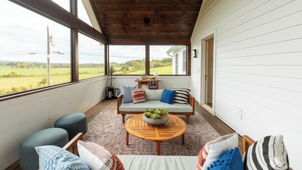 A covered porch with a wooden ceiling, a light-colored sofa with cushions, a round wooden table with a bowl of greenery, and a view of a green field through large windows.
