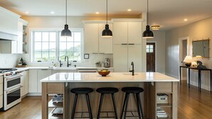 A modern kitchen with white cabinetry, a large island with stools, pendant lighting, and a view of the outdoors through a large window.
