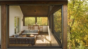A covered outdoor seating area with a sofa, chairs, and a table, overlooking a forested area.