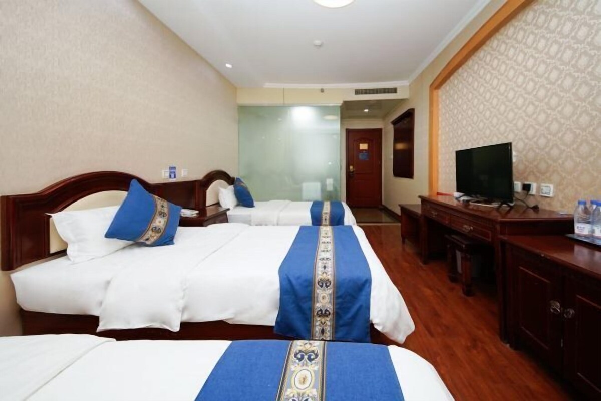 A hotel room with three beds, each with a blue and white bedspread, a television on a wooden desk, and a patterned wall.
