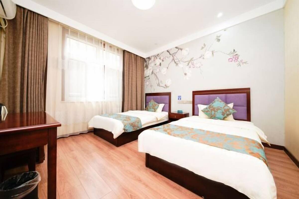 A hotel room with two beds, a wooden desk, a trash bin, and a window with curtains.