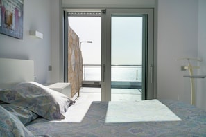 A bedroom with a bed, a nightstand, a wall-mounted lamp, and a view of the sea through an open door.
