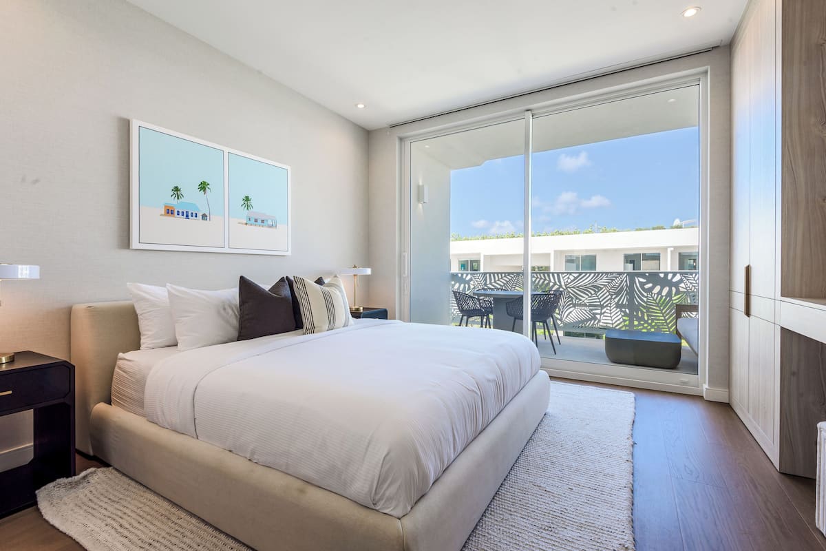 A modern bedroom with a large bed, a nightstand, a wall-mounted TV, and a sliding glass door leading to an outdoor area.