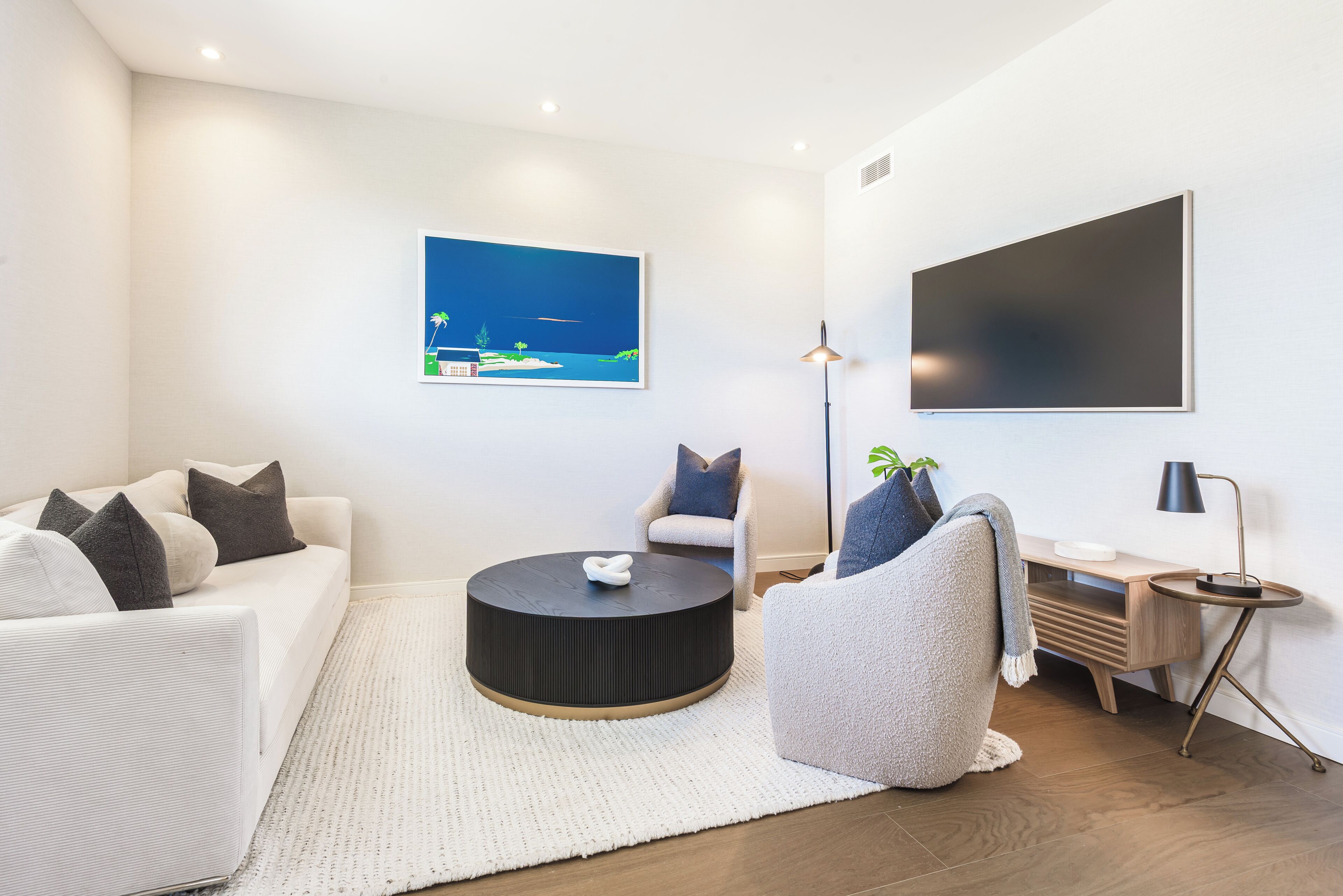 A modern living room with a white sofa, a round coffee table, a side table, a lamp, and a painting on the wall.