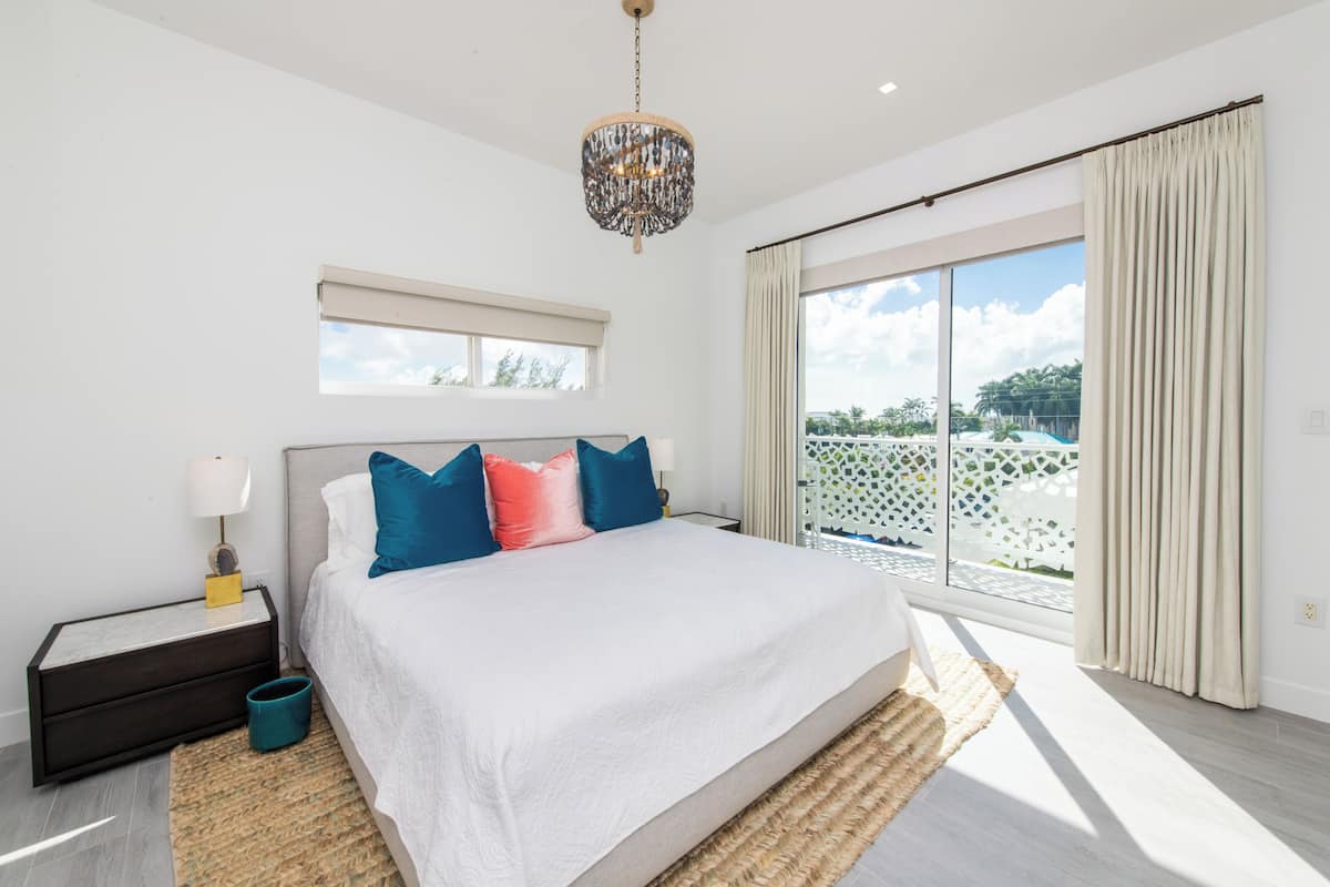 A bedroom with a large bed, a nightstand, a lamp, a window with curtains, and a view of the outdoors.