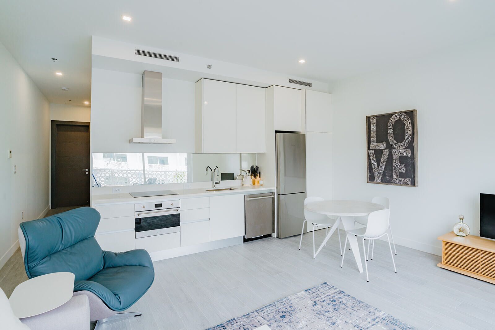 A modern kitchen with white cabinets, a stainless steel refrigerator, and a built-in oven. There is a dining area with a white table and chairs, and a blue sofa. A large 'LOVE' sign is visible on the wall.