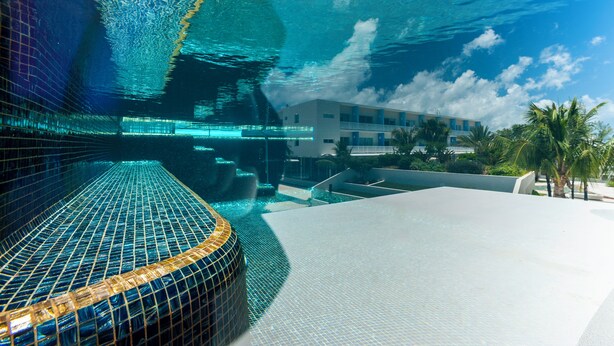 An underwater view of a swimming pool with a modern design, clear water, and a building in the background. - The Grove (Seven Mile Beach)