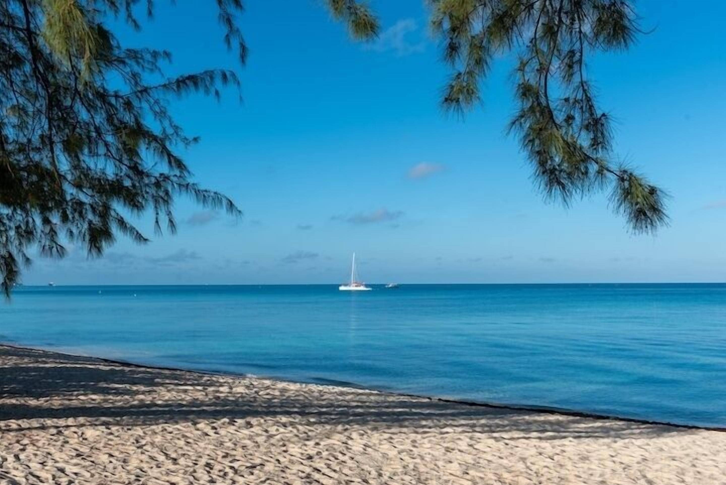 A beach with clear blue water, a distant sailboat, and a sandy shore.