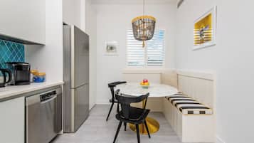 A compact kitchen with a built-in dishwasher, a refrigerator, a round dining table with chairs, and a bench with a striped cushion.