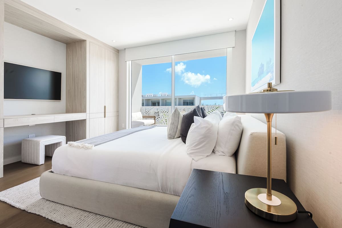 A modern hotel room with a large bed, a flat-screen TV, a bedside table with a lamp, and a view of the city through large windows.