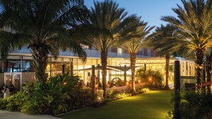 A hotel courtyard with palm trees, a grassy area, and a building with large windows. - The Grove (Seven Mile Beach)