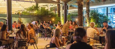 An outdoor dining area with tables and chairs, surrounded by palm trees and greenery.