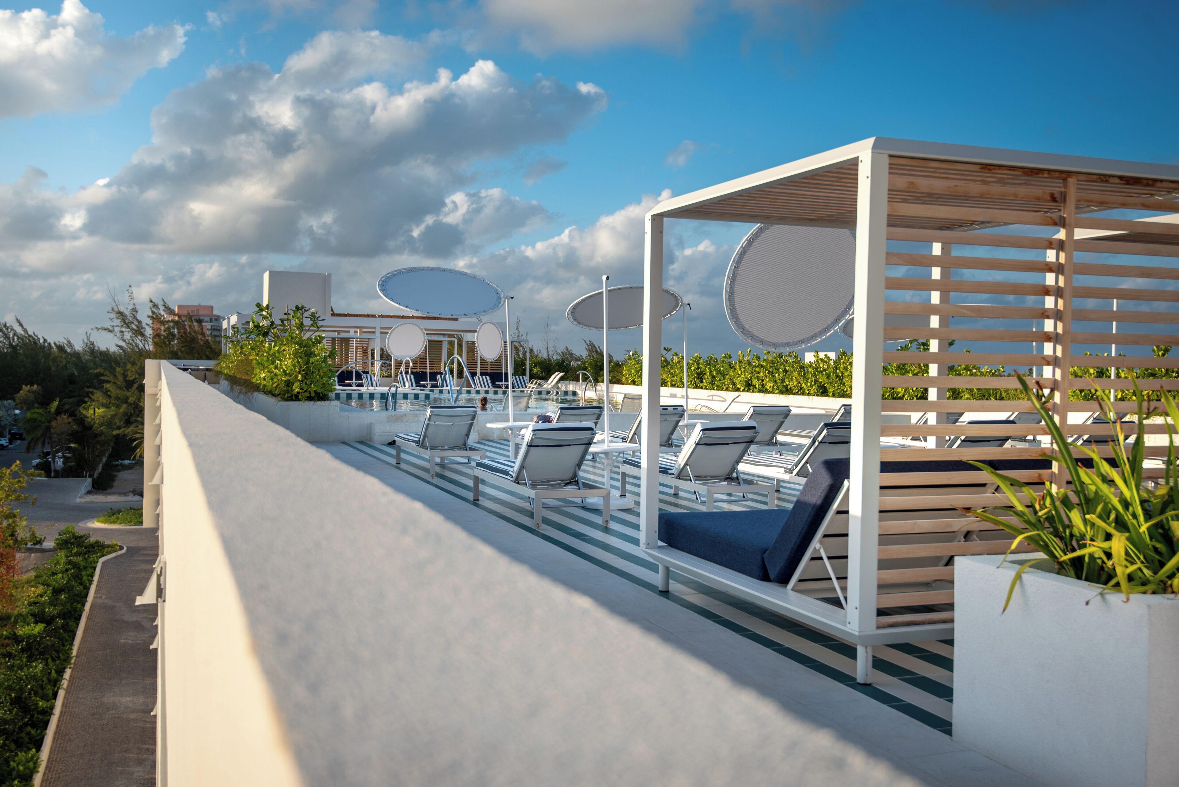 A rooftop pool area with lounge chairs and umbrellas, surrounded by greenery and a clear sky.