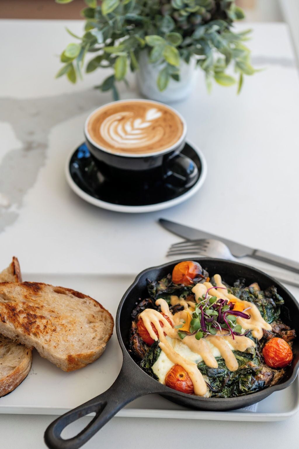 A breakfast setting with a skillet of eggs and vegetables, toast, and a cup of coffee on a table.