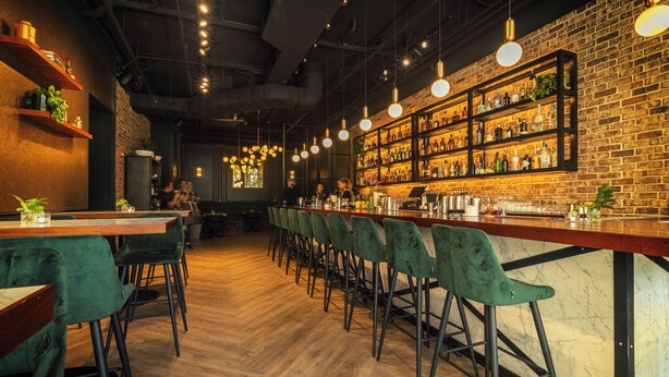 A bar area with wooden floors, green upholstered chairs, and a brick wall with shelves of bottles. - The Grove (Seven Mile Beach)