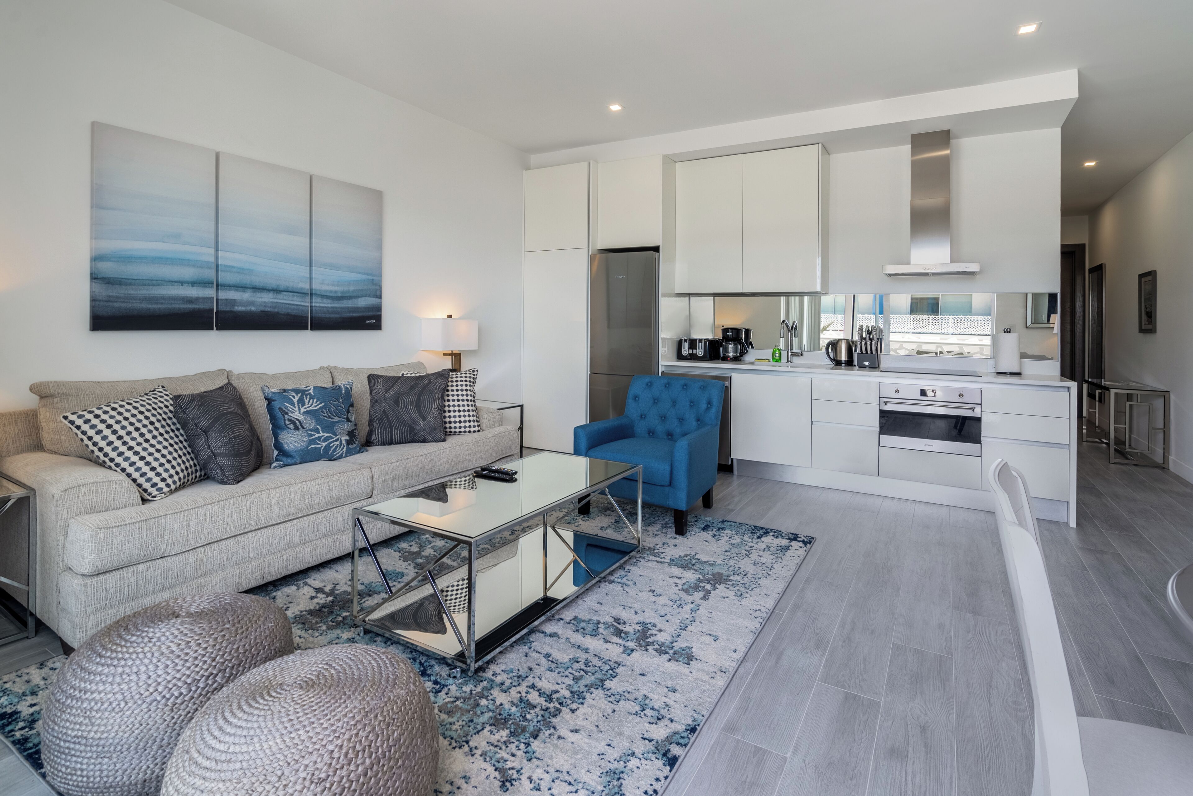 A modern living room with a sofa, coffee table, and blue armchair. A kitchen with white cabinets and appliances is visible in the background.