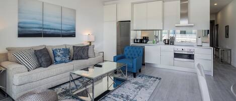 A modern living room with a sofa, coffee table, and blue armchair. A kitchen with white cabinets and appliances is visible in the background.