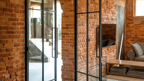 A room with exposed brick walls, a sliding glass door leading to an outdoor area, a sofa, and a small table with folded items.