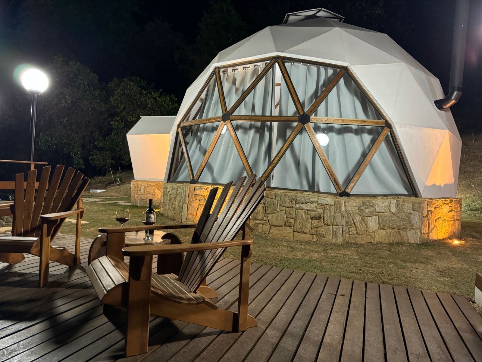 Une structure en forme de dôme géodésique, dotée d’une base en pierre, d’une terrasse en bois et d’un espace extérieur aménagé avec des chaises et une table.