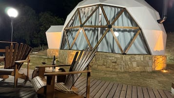 Une structure en forme de dôme géodésique, dotée d’une base en pierre, d’une terrasse en bois et d’un espace extérieur aménagé avec des chaises et une table.