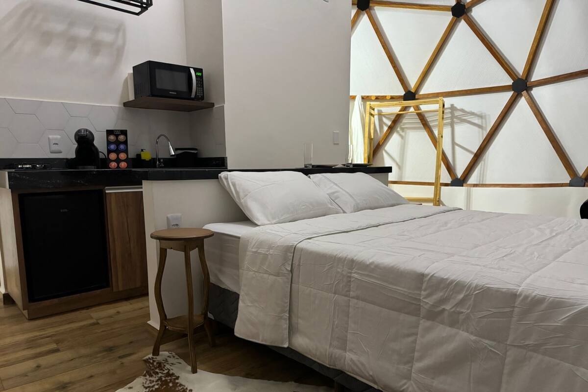 A compact hotel room with a kitchenette, a bed with white bedding, a small stool, and a cowhide rug.