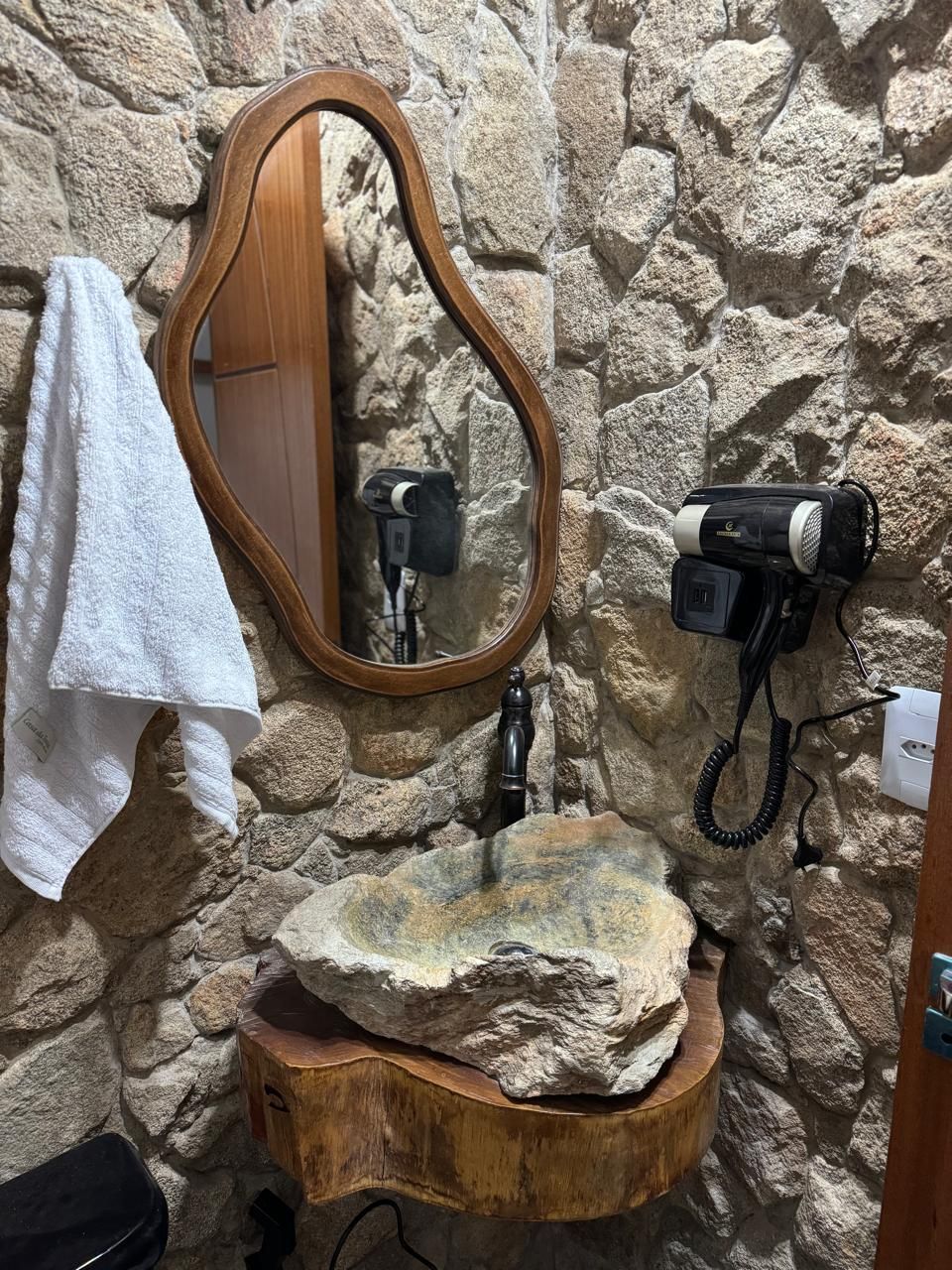 Une salle de bain avec un mur en pierre, un meuble-lavabo en bois avec un évier, un miroir et un sèche-cheveux posé sur un support mural.