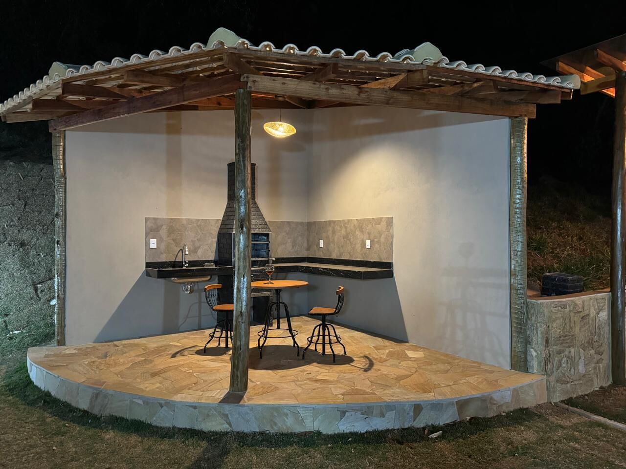 An outdoor patio with a stone floor, a wooden pergola, a built-in grill, and a seating area with stools.