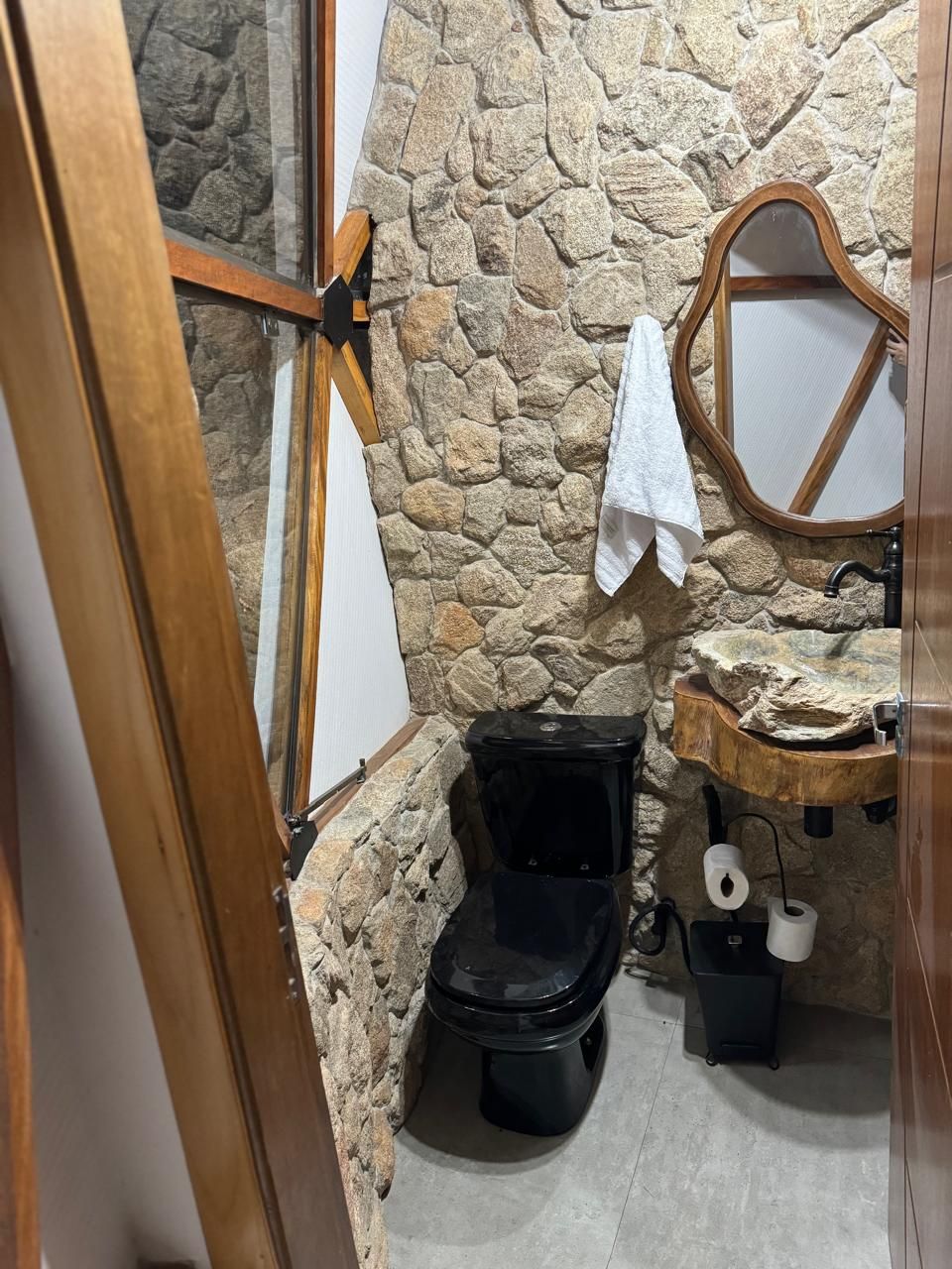 A bathroom with a stone wall, a wooden door, a black toilet, a wooden sink, and a mirror.