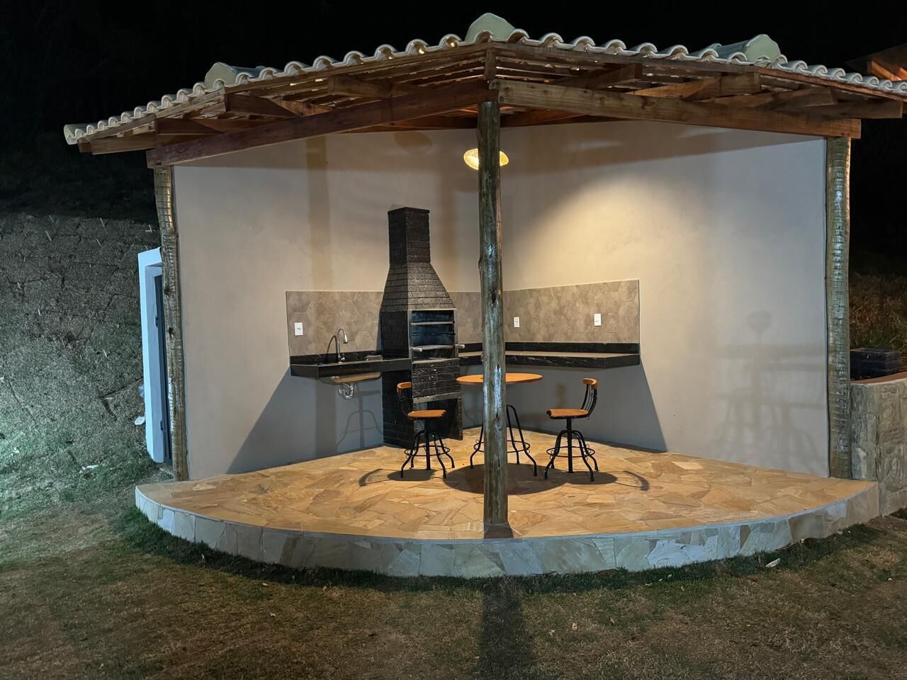 Un patio al aire libre con suelo de losas de piedra, una pérgola de madera, una parrilla empotrada y dos sillas.