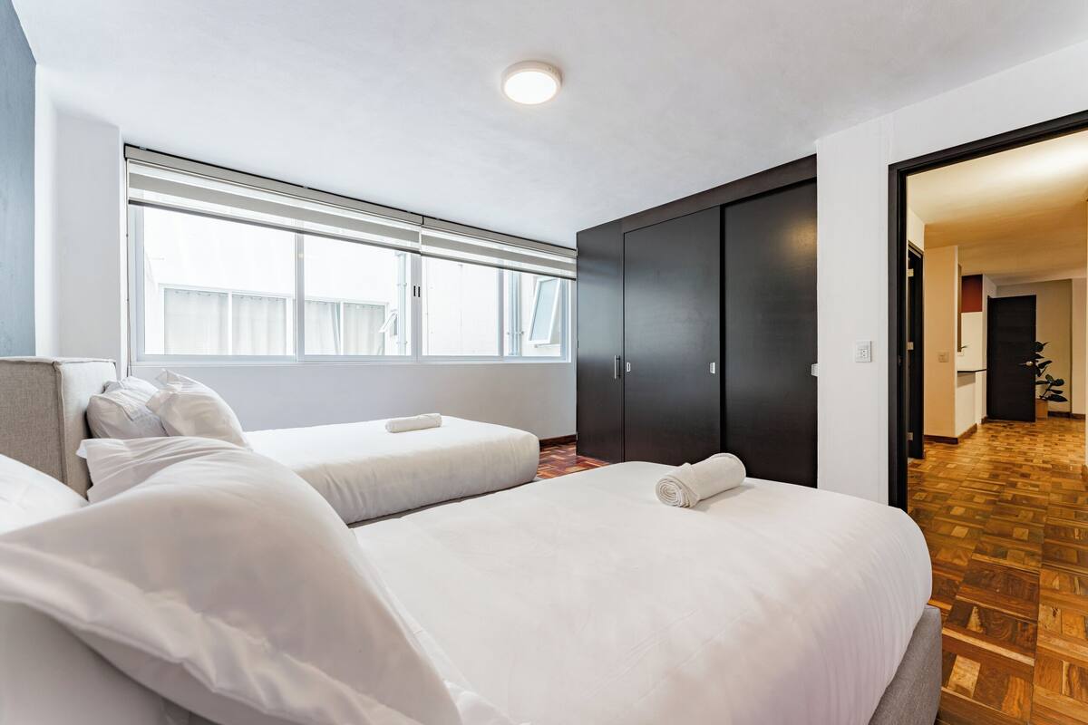 A hotel room with two beds, a large window, a dark wardrobe, and a wooden floor.