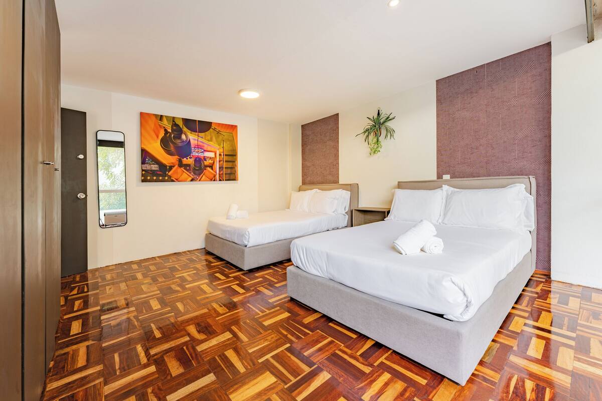 A hotel room with two beds, a wooden parquet floor, a painting on the wall, and a potted plant.