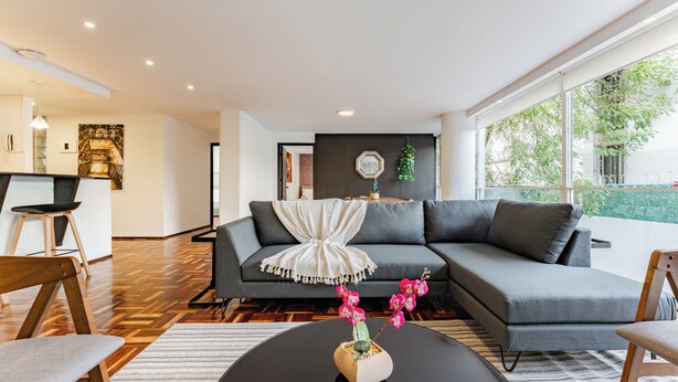 A modern living room with a grey sofa, a round coffee table with a vase of flowers, and a large window offering a view of greenery.