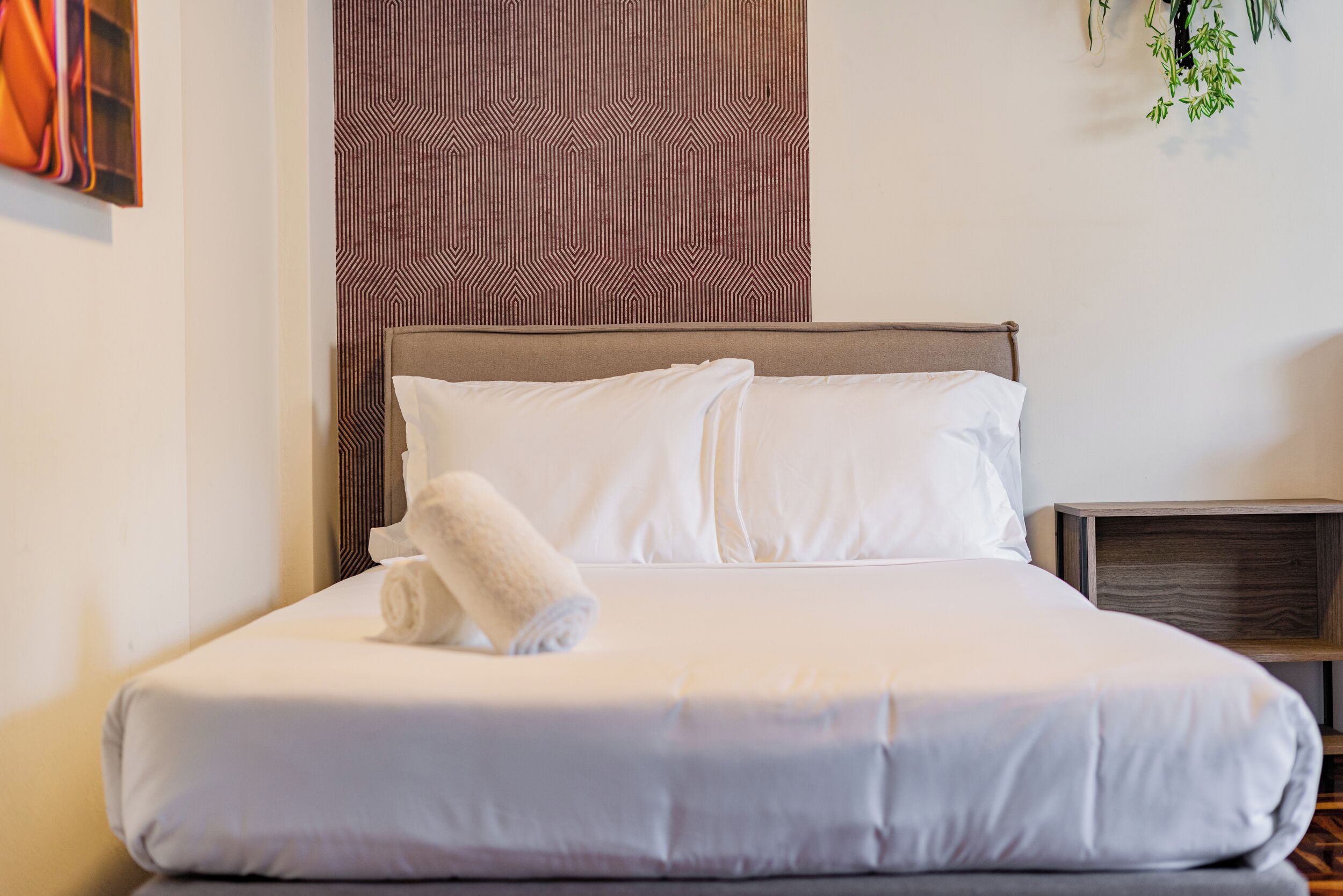 A neatly made bed with white linens and a rolled towel, a patterned wall behind, and a small wooden nightstand with a lamp.