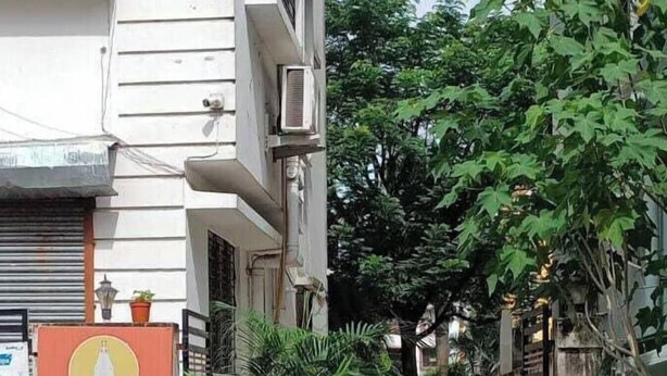 Um beco estreito entre dois edifícios, com aparelhos de ar condicionado fixados nas paredes. Uma placa indica 'Shree Residency Guest House', um guest house com quartos clássicos e deluxe.