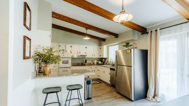 A modern kitchen with white cabinets, a stainless steel refrigerator, wooden beams, and a bar stool.
