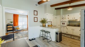 A modern kitchen with white cabinets, a stainless steel refrigerator, and a wooden ceiling. There are two bar stools and a small trash bin.