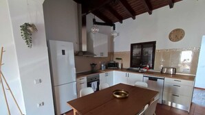A compact kitchen with white cabinets, a wooden dining table, and a hanging plant. - Casa da Pedra (Pedrogão)