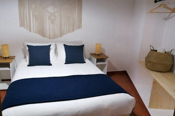 A neatly made bed with a white and blue color scheme, two bedside tables with lamps, a hanging macrame wall decoration, and a wooden bench with a woven bag. - Casa da Pedra (Pedrogão)