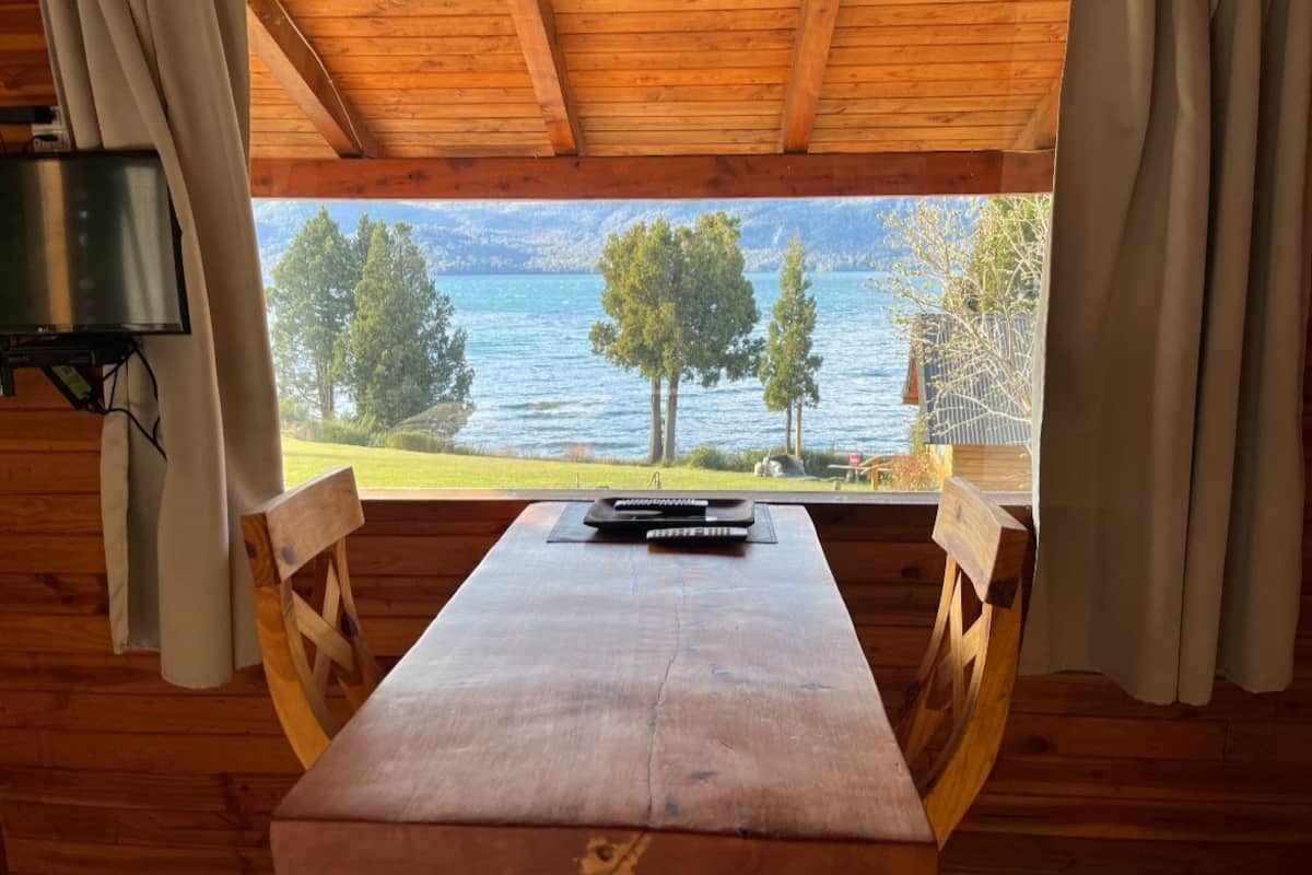 A wooden cabin with a table and chairs, a television, and a view of a lake and trees.