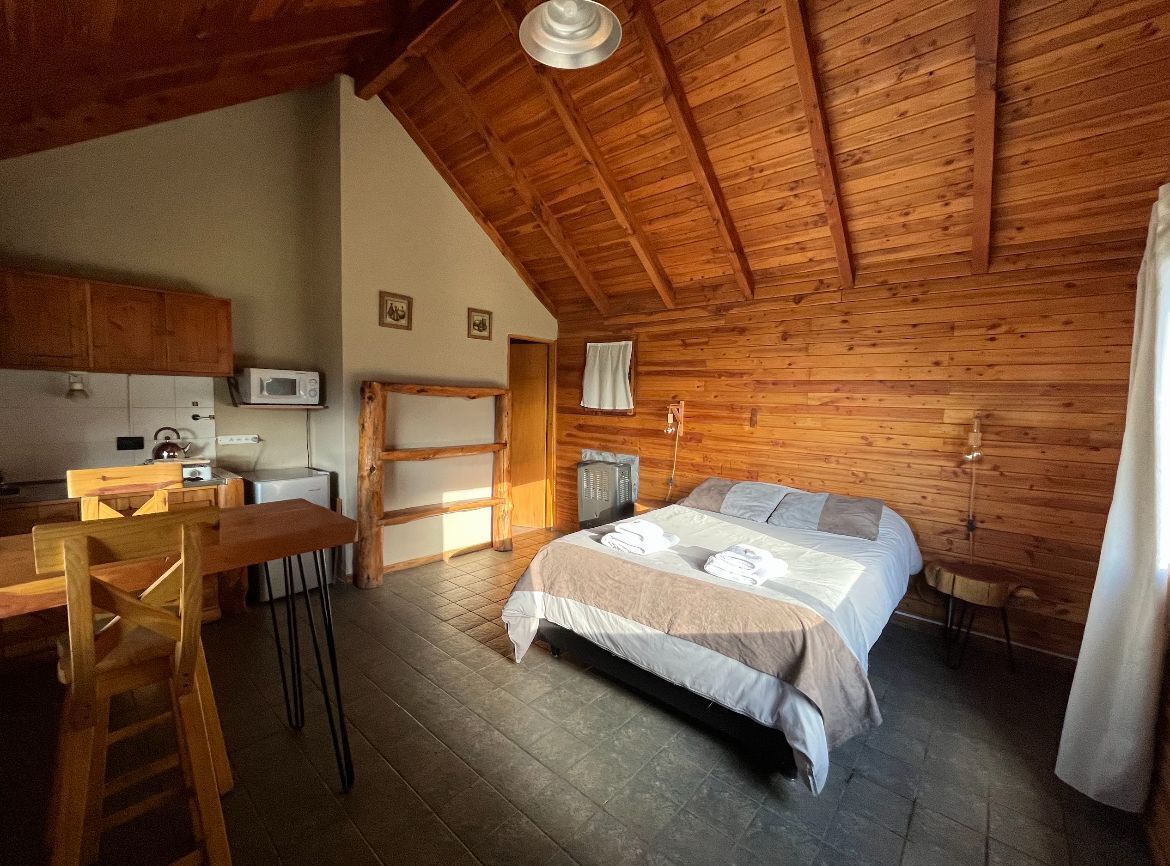 Una habitación con techo de madera, una cama con edredón blanco y gris, una mesa de comedor de madera con sillas, un microondas y una pequeña kitchenette.
