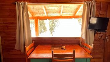 Una baita in legno con un tavolo e sedie, una TV a muro e una finestra con tende.